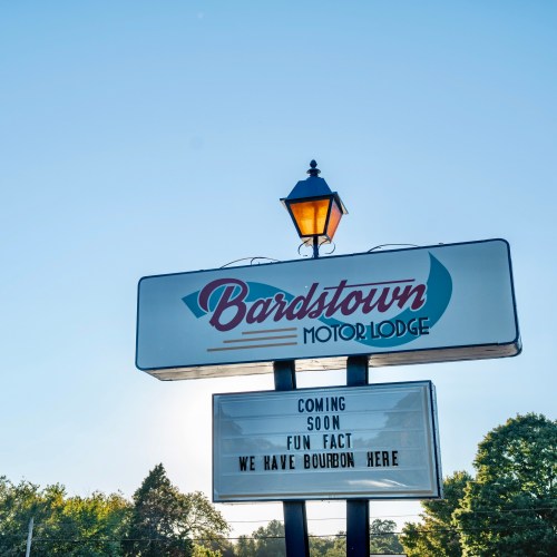 Sign for Bardstown Motor Lodge with text: "Coming Soon Fun Fact We Have Bourbon." Bright sky in the background.