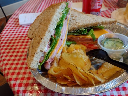 A sandwich with layers of meat and cheese, served with potato chips and a small cup of pickles on a checkered tablecloth.