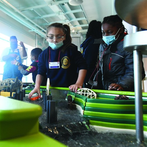Children wearing masks engage with a hands-on exhibit featuring a miniature city model in a bright, interactive space.
