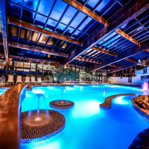 A serene indoor pool area with blue water, fountains, and a wooden ceiling, illuminated by soft lighting.