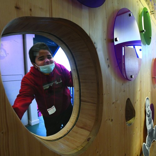 A child in a red jacket smiles while peering through a circular window in a colorful play area.