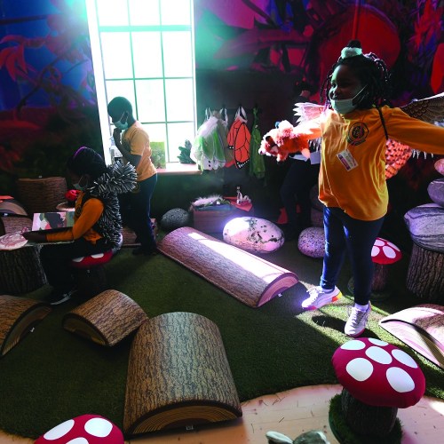Children in colorful outfits engage in activities in a whimsical, nature-themed classroom with logs and mushrooms.