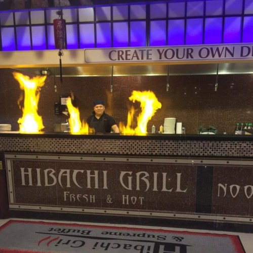 A chef stands behind a hibachi grill, flames rising dramatically as he prepares food in a vibrant restaurant setting.