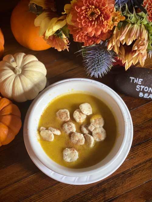 A bowl of yellow soup with dumplings, surrounded by pumpkins and a floral arrangement on a wooden table.