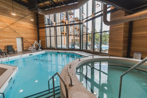 Indoor pool area featuring a large swimming pool and a hot tub, surrounded by large windows and wooden walls.