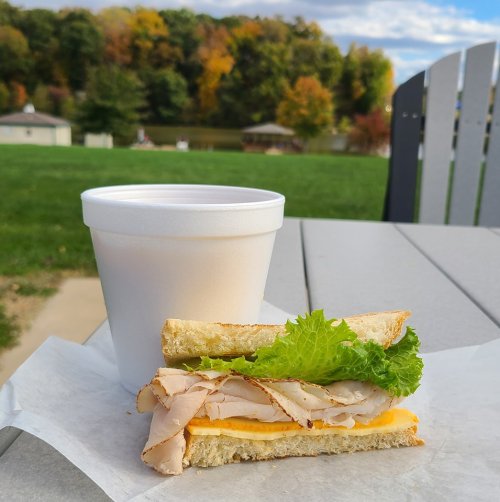 A sandwich with turkey and cheese, topped with lettuce, next to a white cup, set against a scenic outdoor background.