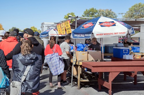 A busy outdoor market with a long line at a hot dog stand under colorful tents. People are chatting and waiting.