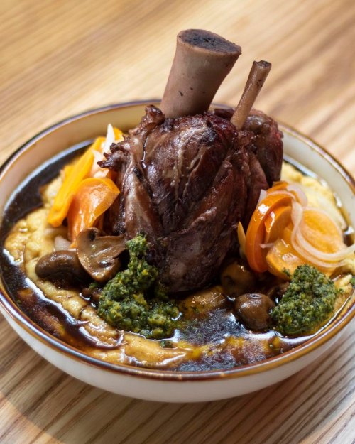 A hearty dish featuring a braised meat shank atop creamy polenta, garnished with vegetables and green sauce.