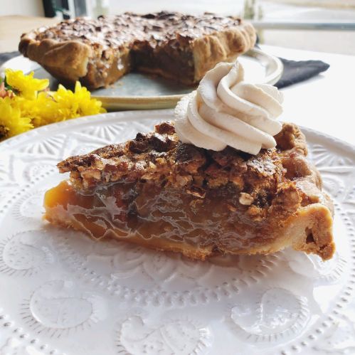 A slice of pie topped with whipped cream, served on a decorative plate with a whole pie and flowers in the background.