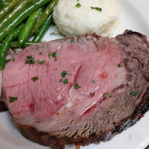 A slice of juicy roast beef with a pink center, served with green beans and a scoop of mashed potatoes, garnished with parsley.
