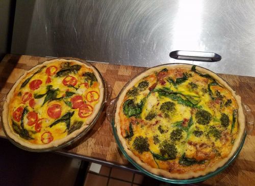 Two quiches on a wooden cutting board: one with tomatoes and spinach, the other with broccoli and spinach.