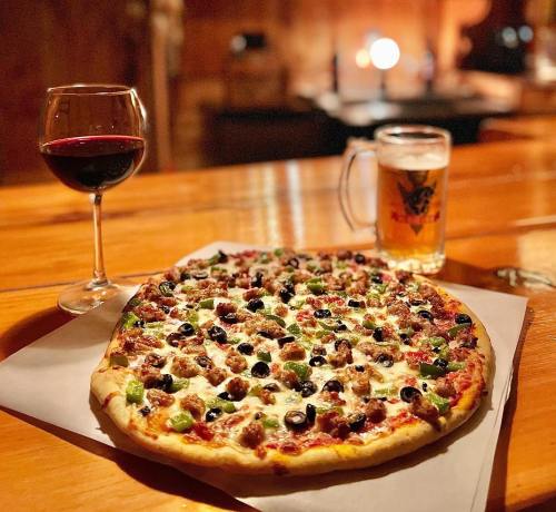 A delicious pizza topped with sausage, olives, and green peppers, accompanied by a glass of red wine and a beer.