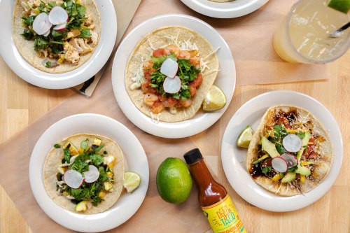 A variety of tacos on plates, topped with fresh ingredients, alongside a lime and a bottle of hot sauce.