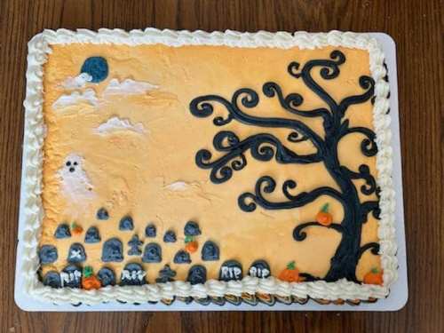 A Halloween-themed cake with an orange background, a spooky tree, tombstones, pumpkins, and a ghost.