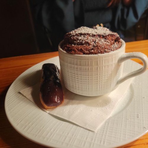 A cup of chocolate soufflé dusted with powdered sugar, next to a chocolate-covered eclair on a white plate.