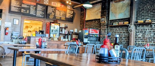 Interior of a casual restaurant featuring wooden tables, condiment holders, a menu board, and a brick wall.