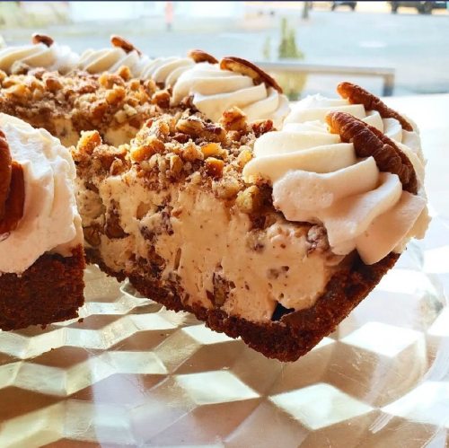 A close-up of a creamy dessert with whipped cream, pecans, and a crumbly crust, partially sliced on a glass plate.
