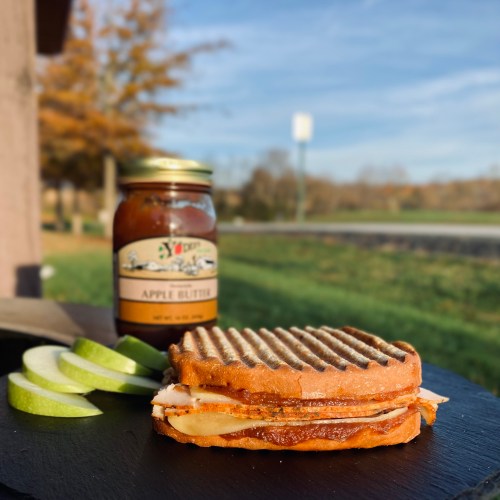 A grilled sandwich with apple slices and a jar of apple butter on a table, set against a scenic outdoor background.