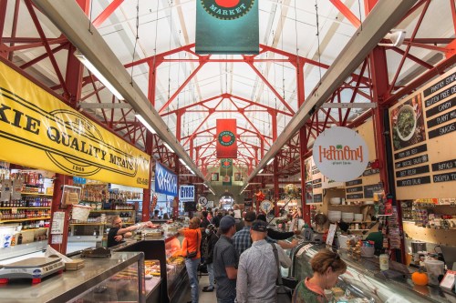 A bustling market interior with colorful vendor signs, shoppers, and a high ceiling adorned with banners.