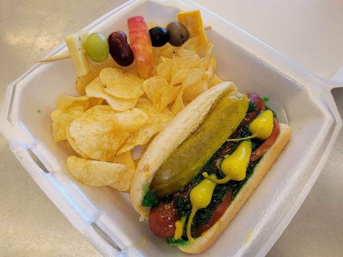 A hot dog with mustard and pickle, served with potato chips and a skewer of fruit and cheese.