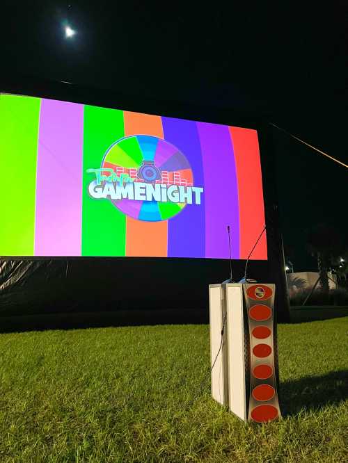 A large outdoor screen displays "GAMENIGHT" with colorful stripes, next to a white speaker on the grass at night.