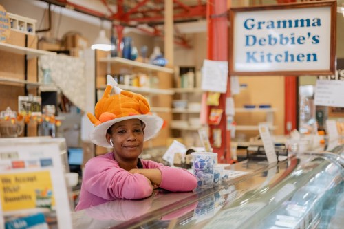 A woman in a colorful hat leans on a counter in a cozy kitchen shop, smiling at the camera.