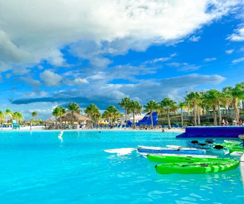 A vibrant beach scene with turquoise water, kayaks lined up, palm trees, and a cloudy blue sky.