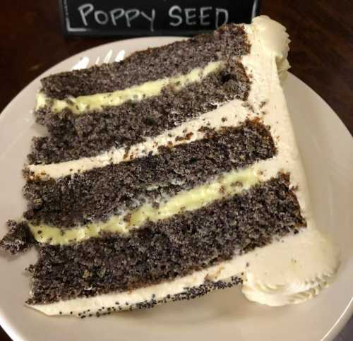 A slice of poppy seed cake with layers of creamy filling and frosted edges on a white plate.