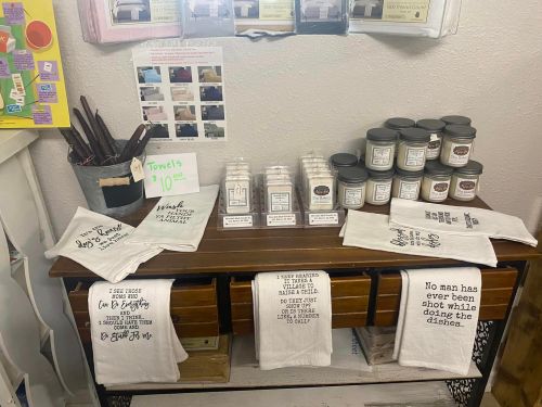A display table with towels, candles, and various items for sale, featuring humorous quotes on the towels.