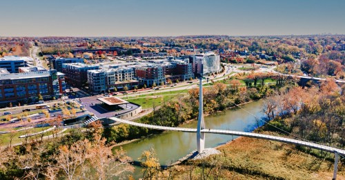 Aerial view of a modern urban area with a bridge over a river, surrounded by trees and buildings in autumn colors.