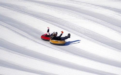 Two people in snow tubes slide down a snowy slope, waving their arms in excitement.