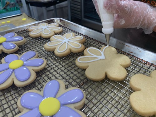 A hand decorates flower-shaped cookies with white icing on a cooling rack, surrounded by colorful icing details.