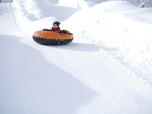 A child in a bright orange snow tube slides down a snowy slope, surrounded by white snowbanks.