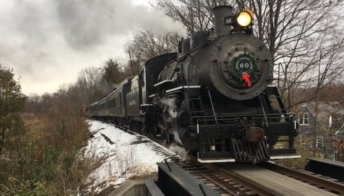 A vintage steam locomotive with a red ribbon travels along a snowy track, surrounded by trees and a cloudy sky.