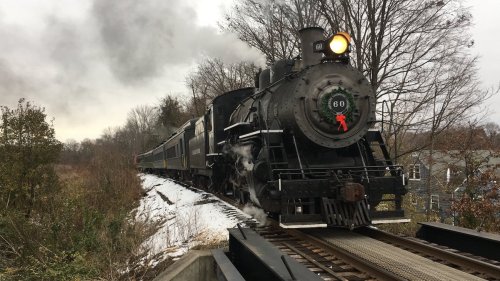A vintage steam locomotive with a red wreath on the front, traveling along a snowy track surrounded by trees.