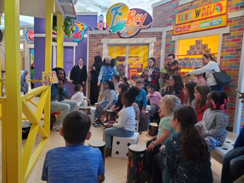 A diverse group of children and adults gather around a performer in a colorful play area, engaging in a music session.