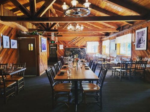 Cozy wooden restaurant interior with tables, chairs, and rustic decor, featuring a stone fireplace and warm lighting.