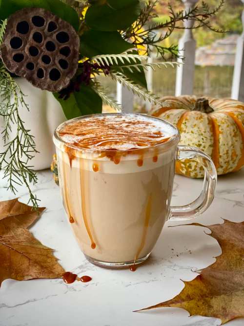 A glass mug filled with creamy coffee topped with caramel drizzle, surrounded by autumn leaves and decorative pumpkins.