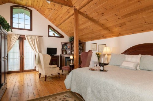 Cozy bedroom with wooden beams, a large bed, armchairs, and a small table with champagne, featuring natural light.