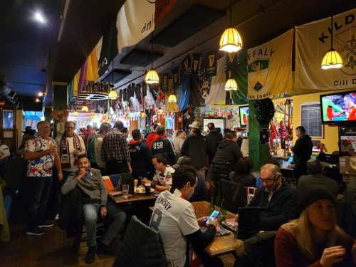 A lively pub scene with patrons enjoying drinks, surrounded by flags and sports memorabilia, watching a game on TV.