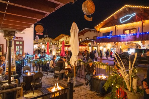 A festive outdoor dining area at night, with people gathered around fire pits and decorated buildings in the background.
