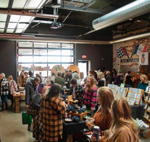 A bustling indoor market with people browsing tables filled with crafts and goods in a lively atmosphere.