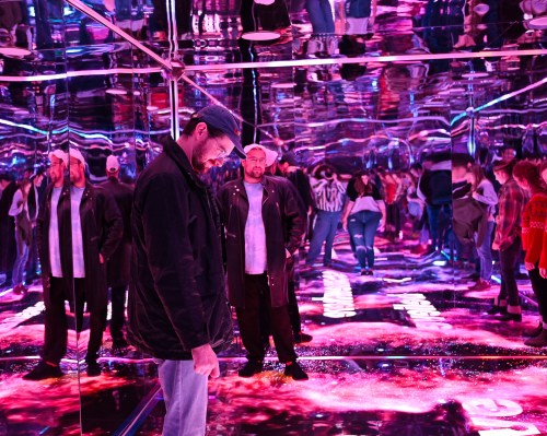 A mirrored room filled with people, reflecting vibrant pink and purple lights, creating a surreal atmosphere.