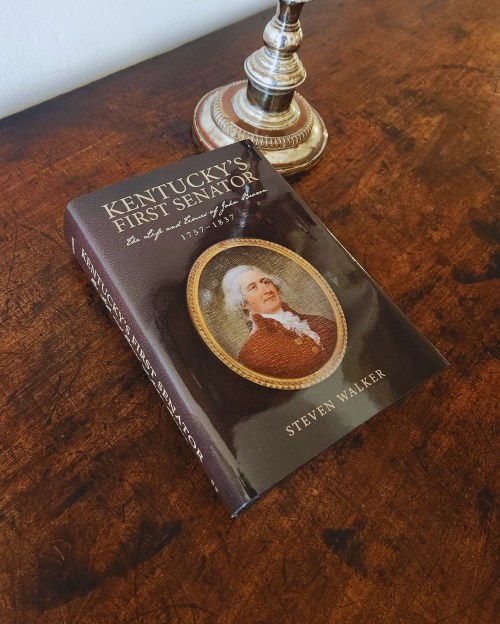 A book titled "Kentucky's First Senator" by Steven Walker, resting on a wooden surface next to a decorative lamp.
