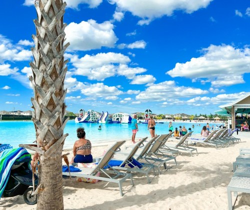 A sunny beach scene with people enjoying a pool, lounge chairs, and inflatable water slides under a blue sky.