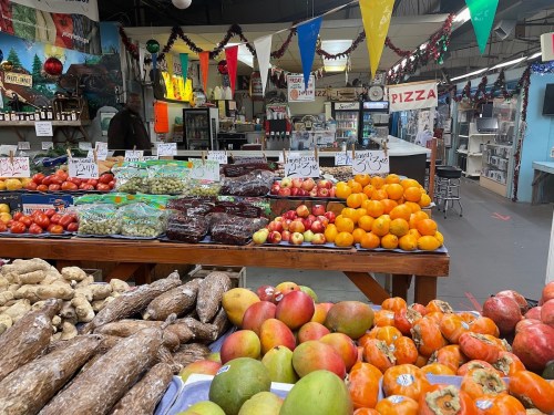 A vibrant market display featuring fresh fruits and vegetables, with colorful banners and a pizza sign in the background.