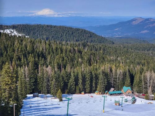 A snowy mountain landscape with ski lifts, a lodge, and dense evergreen trees, with a distant mountain peak in the background.
