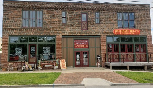 A brick building with large windows, featuring "Railroad Bill's Dining Car" and "Raccoon Farms Trading Co. Antiques" signs.