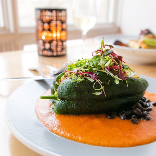 A stuffed pepper on a plate with a vibrant orange sauce, garnished with greens, and a glass of wine in the background.