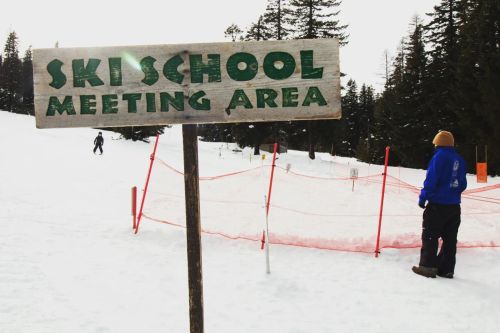 A wooden sign reads "Ski School Meeting Area" with a snowy landscape and a person in ski gear nearby.